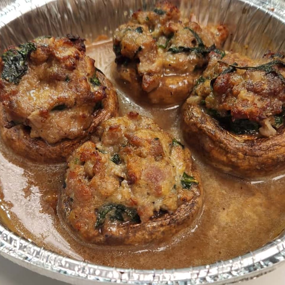 Four stuffed mushrooms with sausage and spinach in a foil container, swimming in a savory sauce.