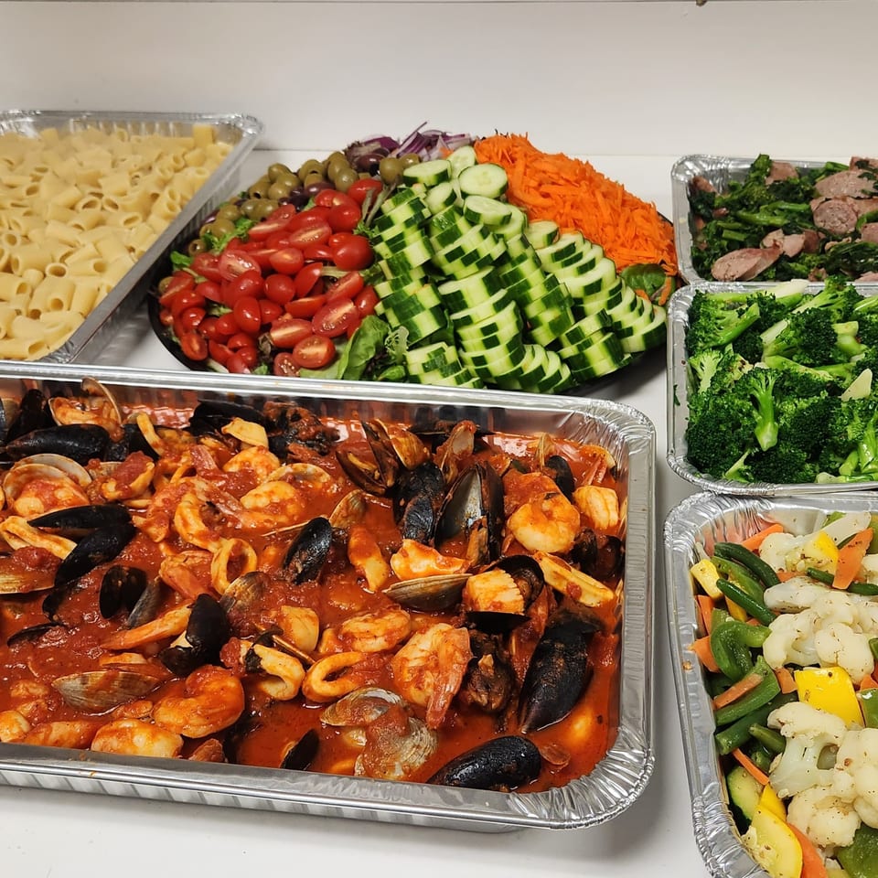 Catering trays of pasta, seafood marinara, salad, broccoli rabe, steamed broccoli, and mixed vegetables.